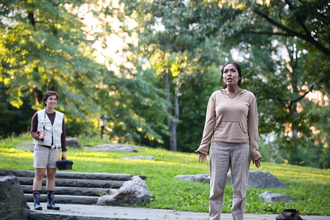 Production photo from Bethel Park Falls of a woman standing while a figure with a fishing pole looks on, outdoors in a park