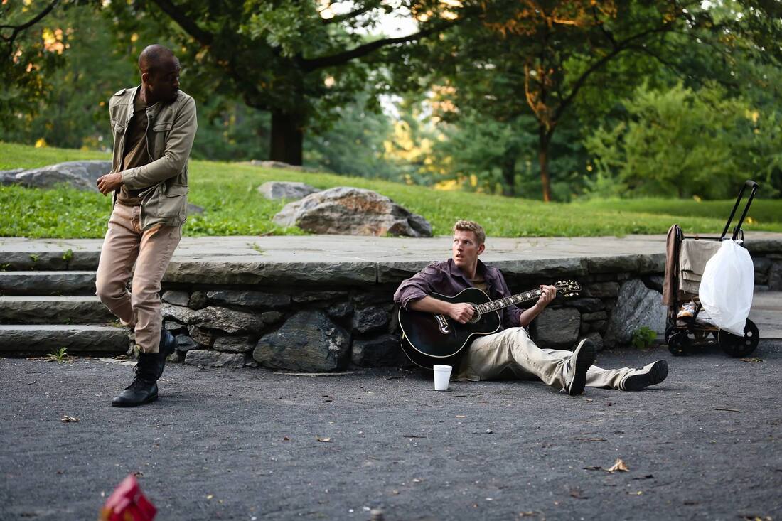 Production photo from Bethel Park Falls of a standing man turning to look at a seated man playing the guitar in the park.