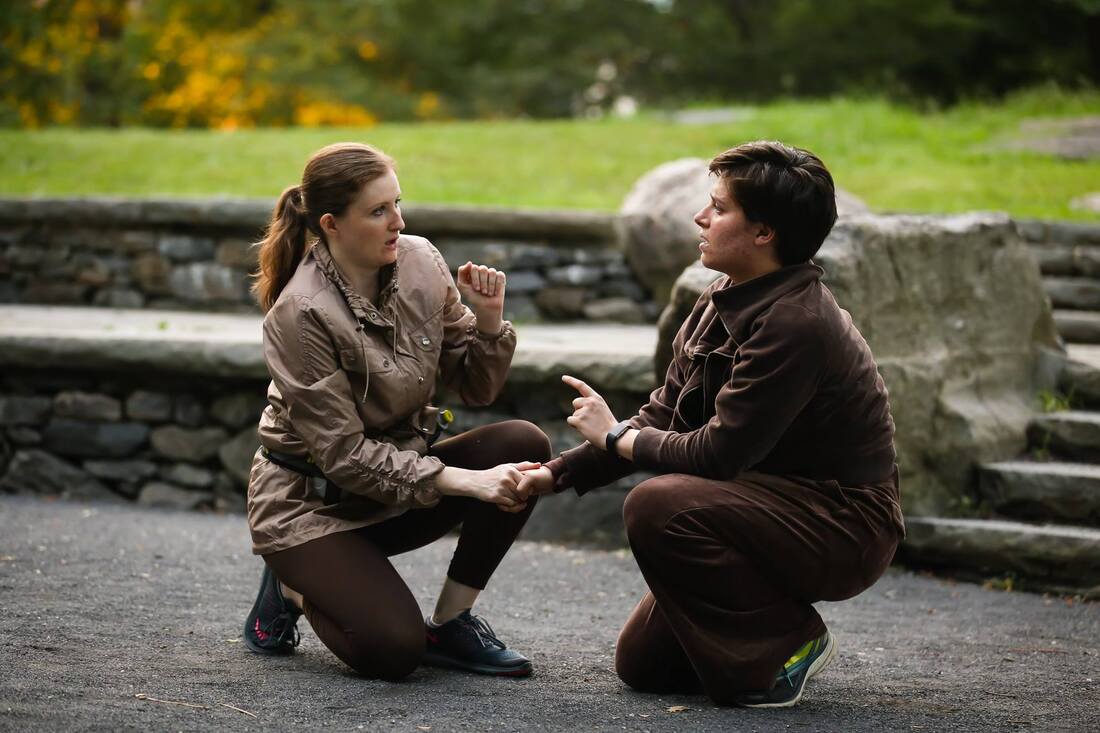 Production photo from Bethel Park Falls of two crouching cast members holding hands and looking at each other, outdoors in a park. 
