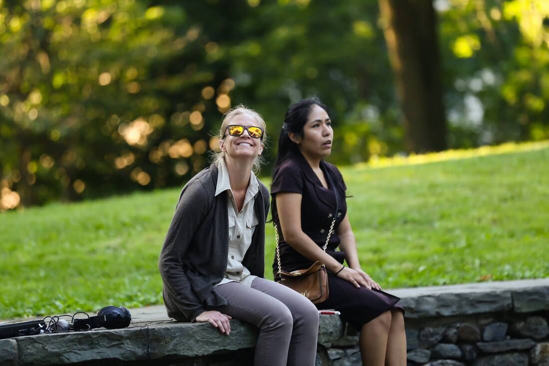 Production photo of two female cast members of Bethel Park Falls sitting on a low wall outdoors in a park.